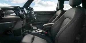 The interior of a MINI Cooper car. The front seats are upholstered in black leather and have a sporty design with bolsters for support. The seats are adjusted to a comfortable driving position, and the headrests are positioned behind the driver and passenger. The dashboard features a large touchscreen display and a digital instrument cluster. The steering wheel is leather-wrapped and has multiple buttons for controls. The center console includes a gear selector, a parking brake, and other buttons. The windows are open, and the sky is visible outside.