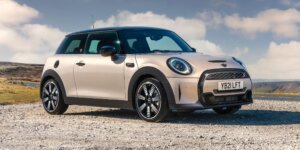 A silver MINI Cooper hatchback car parked on a dirt road with a scenic mountain landscape in the background. The car has a sporty design with black accents and alloy wheels. The license plate on the car is YB21 LFT.