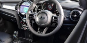 The interior of a MINI Cooper car. The dashboard features a large circular digital display, a leather-wrapped steering wheel with the MINI logo, and a gear selector. The seats are upholstered in black leather with red stitching, and the center console has buttons for various controls.