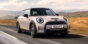 A silver MINI Cooper hatchback car driving on a road with a scenic landscape in the background. The car has a sporty design with black accents and alloy wheels. The license plate on the car is YB21 LFT.