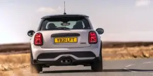 The rear of a silver MINI Cooper hatchback car driving on a road with a scenic landscape in the background. The car has a sporty design with black accents and alloy wheels. The license plate on the car is YB21 LFT.