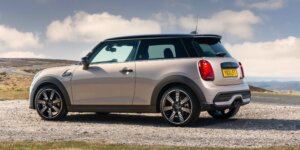 A gray MINI Cooper Hatchback parked on a dirt road with a scenic mountain backdrop. The car has black alloy wheels, a black roof, and a sporty design. The license plate on the car is YB21 LFT.