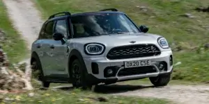 A white MINI Cooper Countryman SUV driving on a dirt road with a scenic landscape in the background. The car has a sporty design with black accents and alloy wheels. The license plate on the car is OX20 CSE.