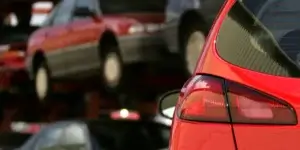 A close-up of the rear of a red car in a junkyard. The car's taillight is visible, and there are other cars partially visible in the background.