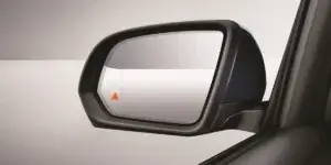 A close-up of a side mirror on a car. The mirror is black with a chrome trim and has a blind spot monitoring system activated, indicated by a small red triangle in the lower corner.